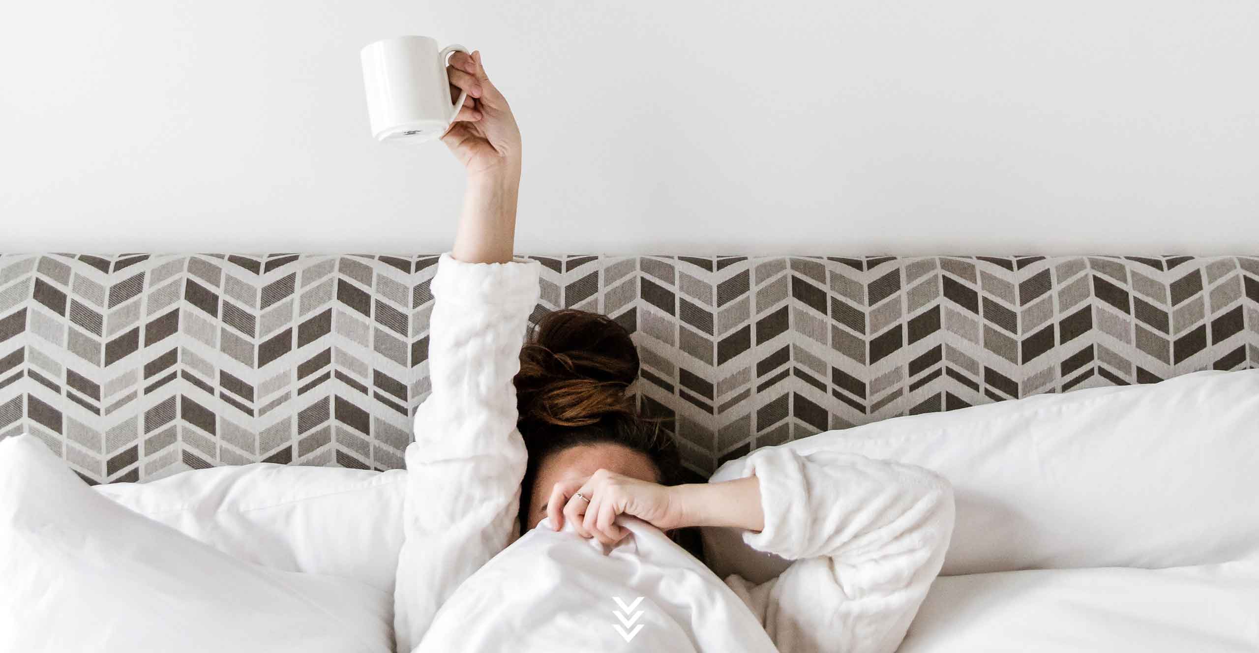 woman in bed with coffee cup covering half her face with blanket