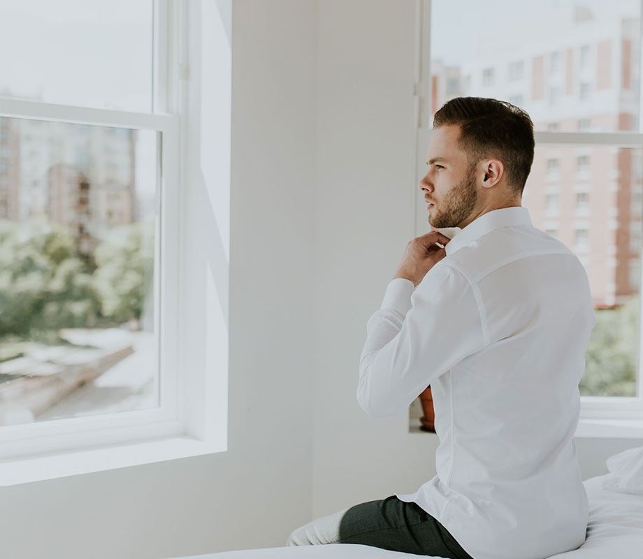 young man in white dress shirt sitting on bed looking out